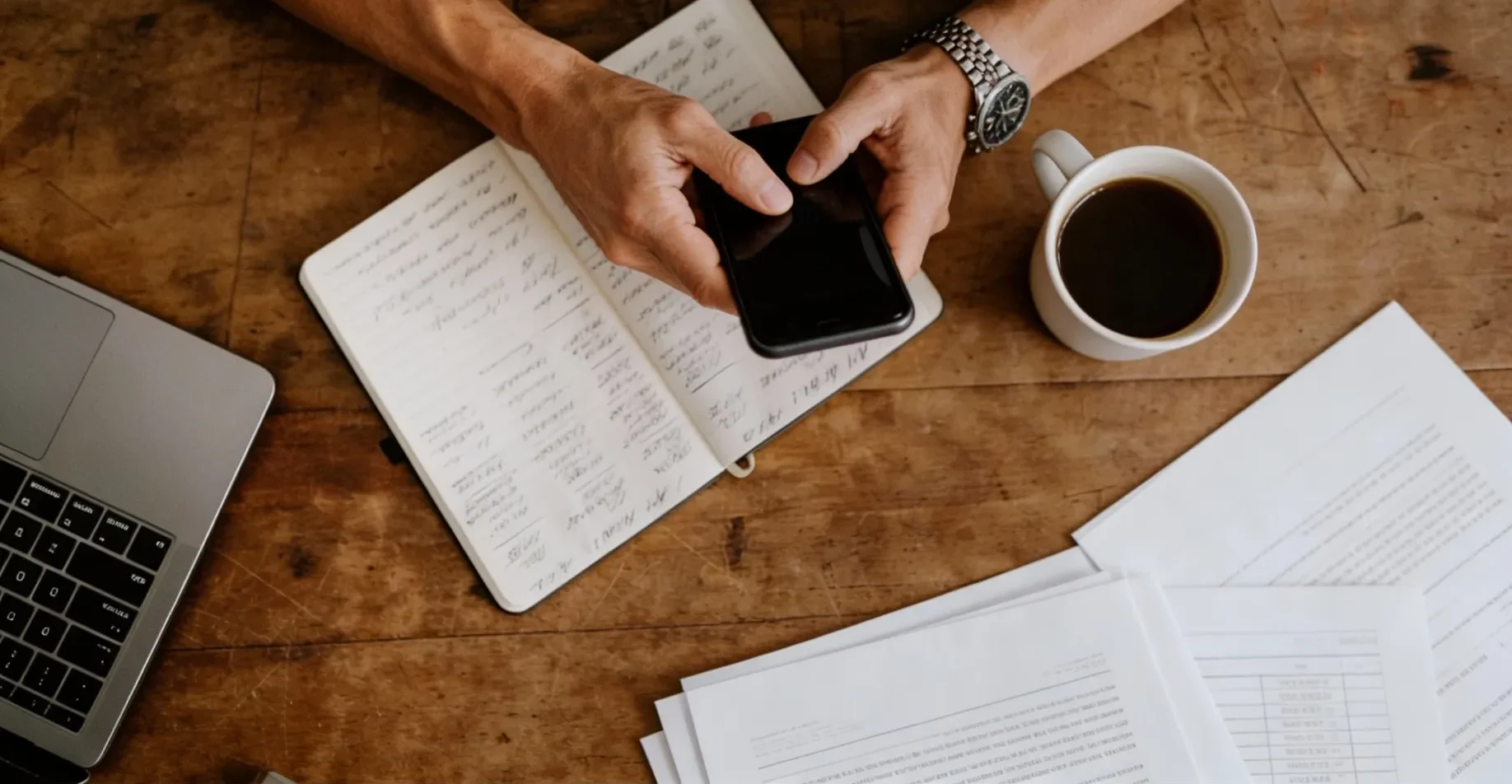 A person holds a smartphone above a notebook, laptop, and a cup of coffee on a wooden table.